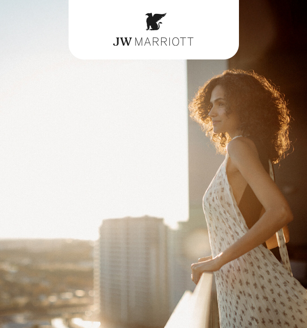 A woman with curly hair stands on a balcony, wearing a light-colored dress, looking out at a cityscape during sunset. The JW Marriott logo is visible at the top.