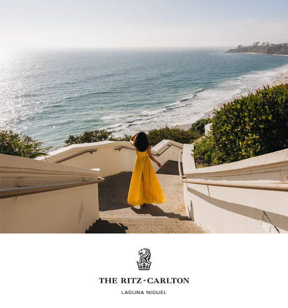 A woman in a yellow dress walking down steps towards a beach, with waves and greenery in the background. The scene is illuminated by sunlight, creating a vibrant atmosphere. The Ritz-Carlton Laguna Nigel logo is at the bottom.