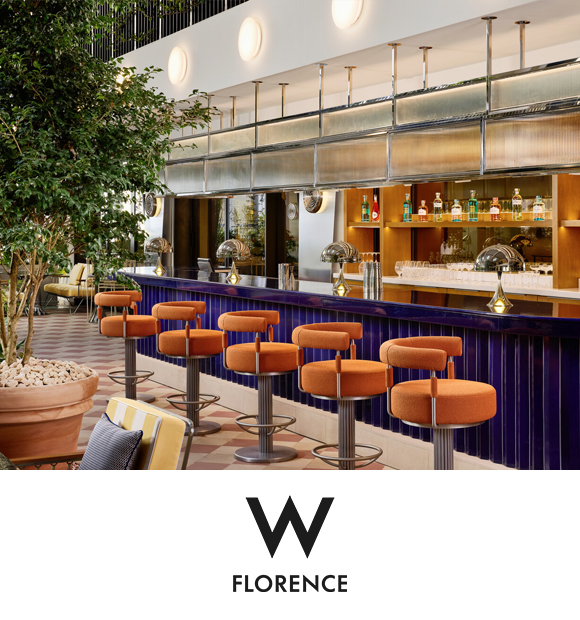 Interior view of a stylish bar at W Florence. Features eight orange bar stools lined up at a modern bar counter, with bar shelves displaying various bottles in the background, accented by greenery and decorative pots. W Florence logo at the bottom.