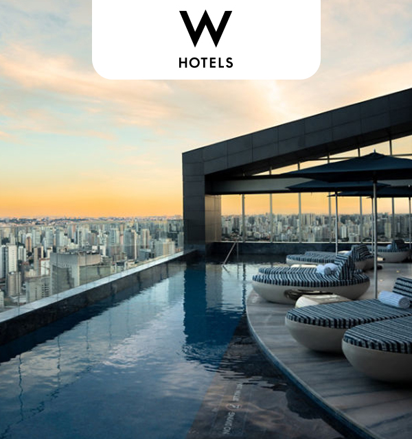 A modern bar area within W Sao Paulo, featuring a backlit onyx bar top and stylish seating. The bar is stocked with various bottles, and large windows reveal a city skyline at night. The W hotels logo is at the top of the image.