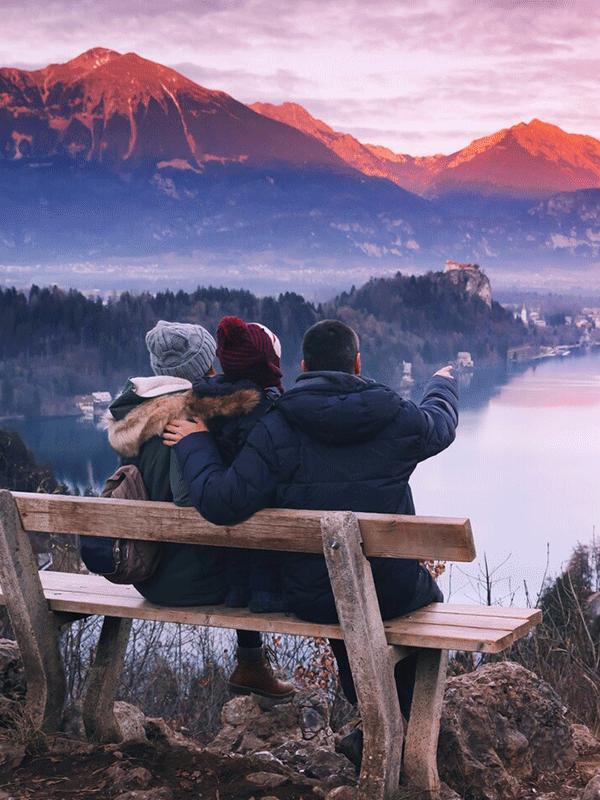 A trio of friends are enjoying the mountainside sunset from a bench.