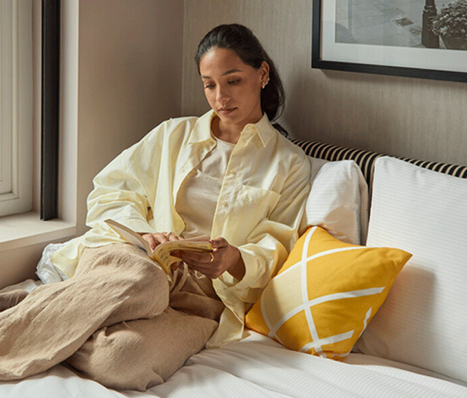A woman of color, dressed in a yellow shirt and relaxed beige pants, is sitting on top of a bed and reading a book by the window.