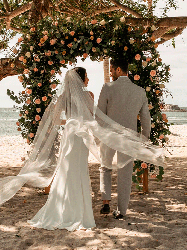 Bride and groom walking down a sandy beach to get married under a floral arch.
