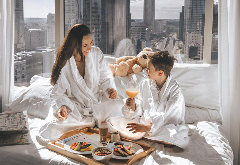 Mother and son having breakfast in bed wearing white robes in front of big window with city skyline in background