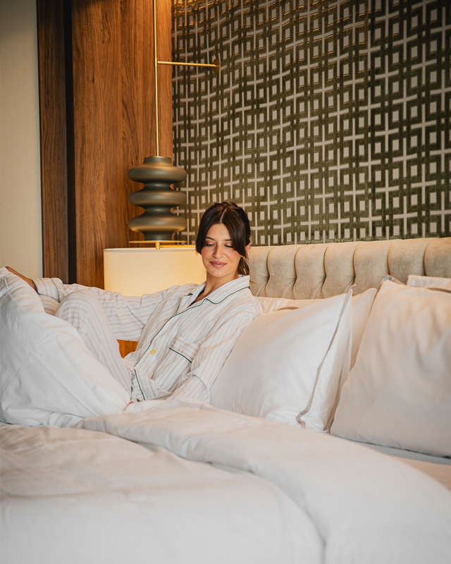 A woman in striped pajamas relaxes on a bed with white pillows and a plush headboard, set against an elegant patterned wall. She appears content and comfortable.