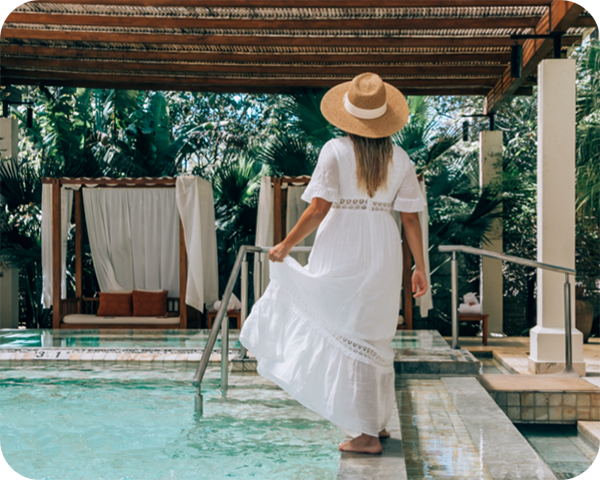 Woman in a white dress and hat walks next to a pool under a pergola with tropical plants.