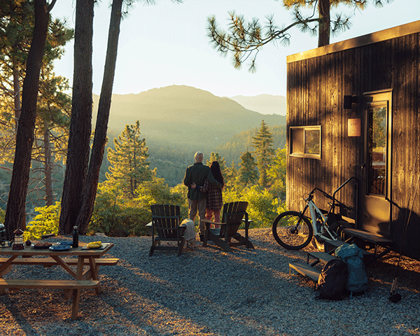 Couple overlooks rolling green mountains at sunset next to a cozy, modern cabin.