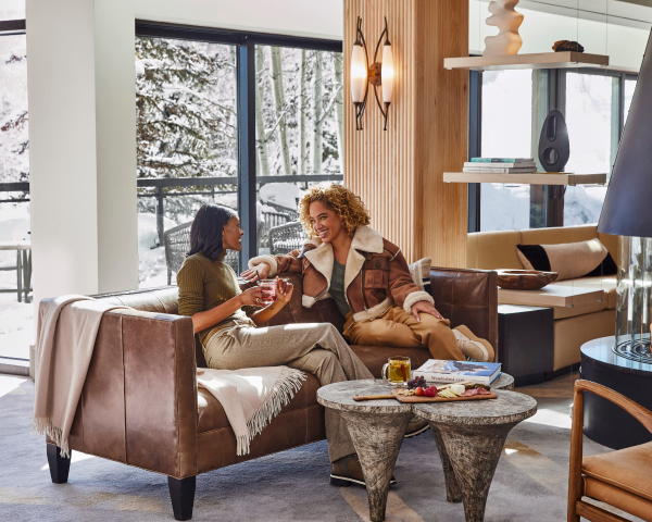 Travelers laugh on a couch in a modern, cozy space with snowy woods outside.