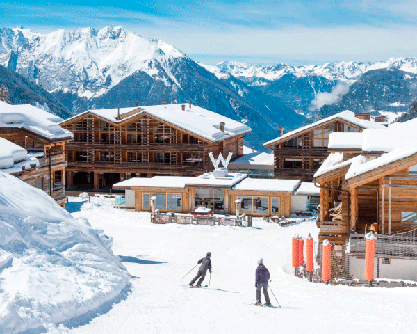 Two skiers at the bottom of a mountain slope, approaching a ski-in/ski-out lodge and resort.