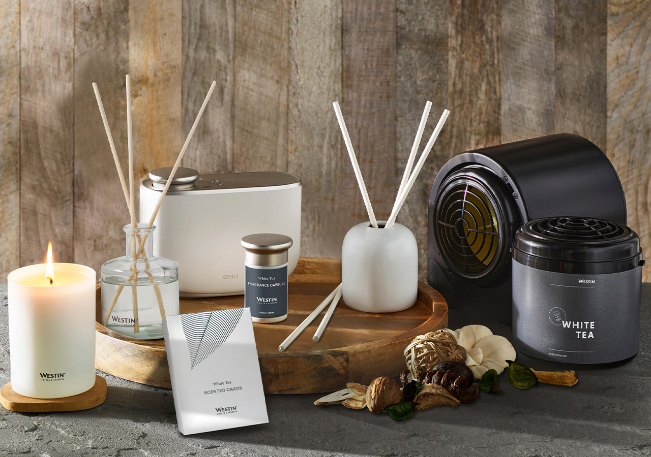 Various fragrances from Westin's White Tea Collection displayed on a gray table and against a wooden wall background. Soft lighting is shining on the products.
