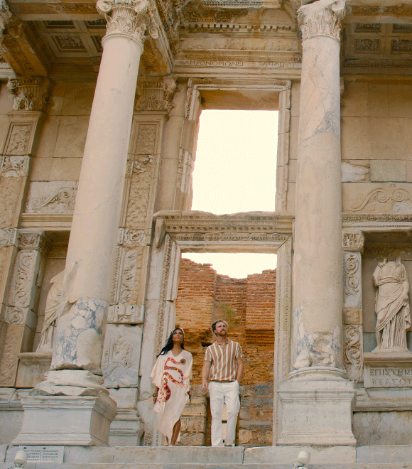 Man and woman standing in front of ancient ruins with ornate columns and sculptures in Athens, Greece, possibly conveying a sense of awe and historical curiosity.