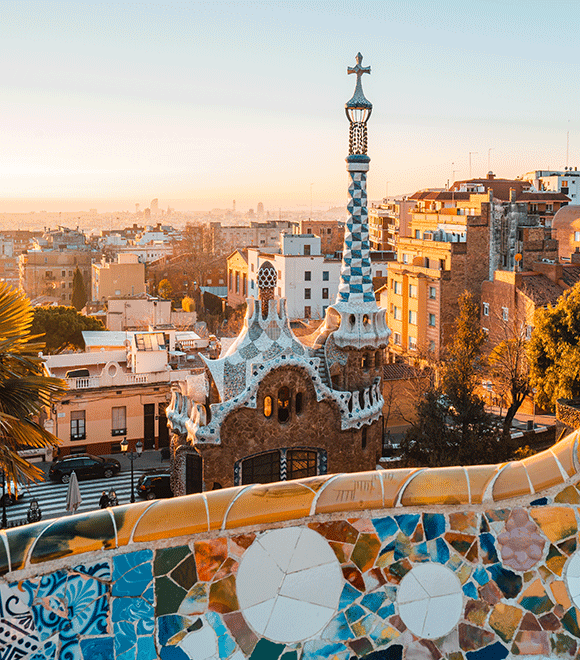 Sunlit view of Park Güell in Barcelona, showcasing Gaudí's colorful mosaic designs in the foreground and cityscape with warm tones in the background.