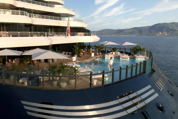 View of Luminara's pool deck with umbrellas, lounge chairs, and plants. Overlooks serene ocean and distant mountains under a clear sky. Relaxing ambiance.