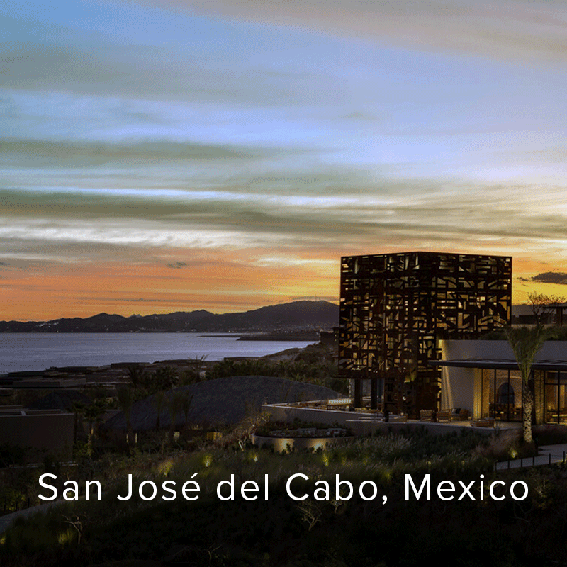 Hotel exterior with a glowing patterned facade overlooks a calm bay and dark mountains at sunset