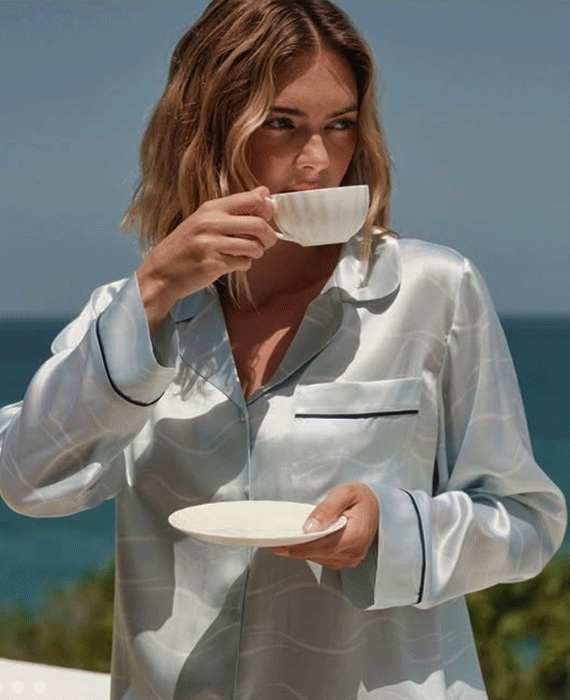 Woman in white satin pajamas sips tea from a cup while holding a saucer outdoors, with blue sea and sky behind her.